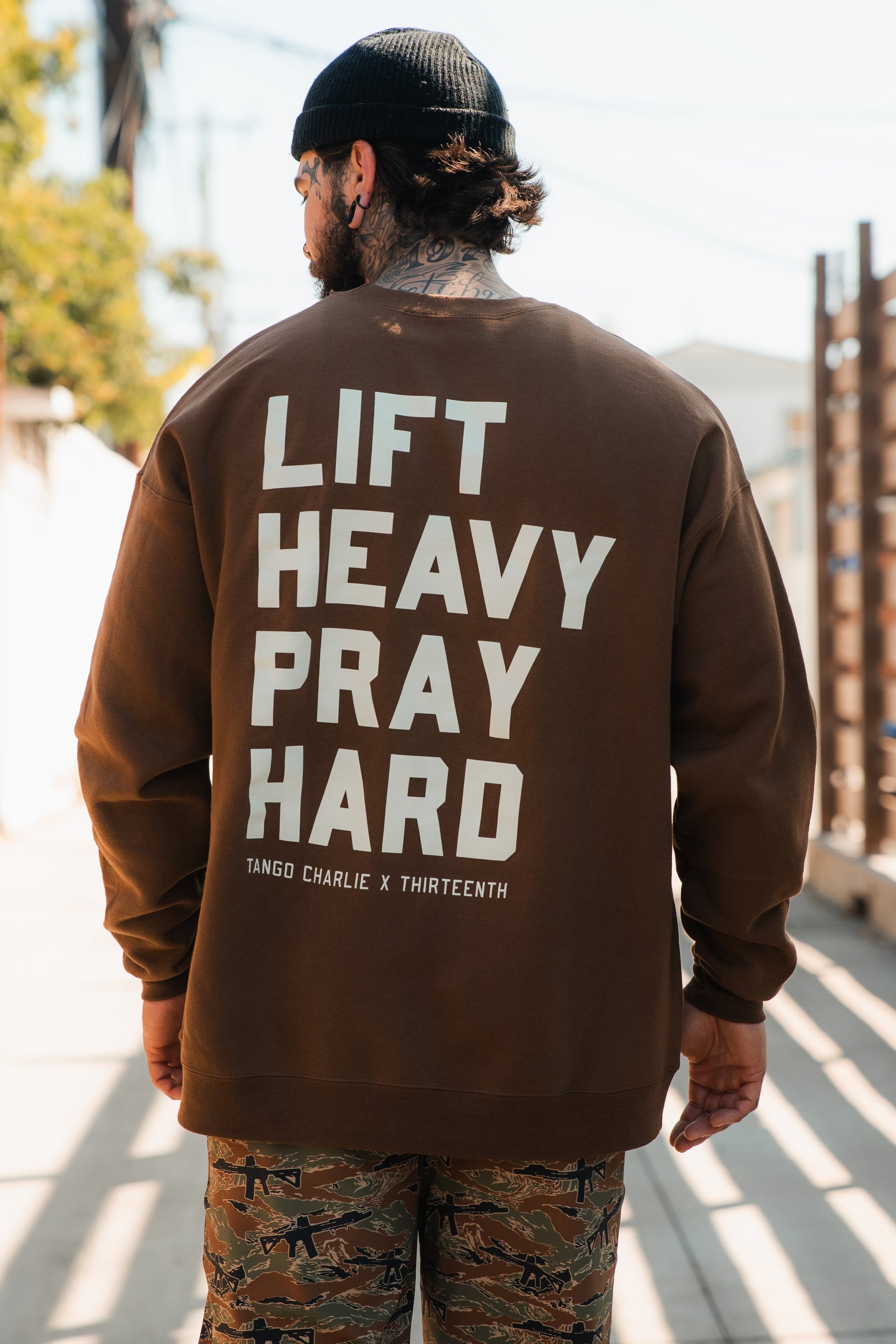 A man stands outdoors, facing away from the camera, wearing the Tango Charlie Apparel "Lift Heavy. Pray Hard." crewneck in brown with bold white text, paired with camo pants. Sunlight and a fence are visible in the background.