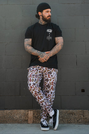 Wearing the "Lift Heavy. Pray Hard." black tee by Tango Charlie Apparel, a tattooed man in a black beanie stands against a gray brick wall, sporting brown and white camo pants and black sneakers—arms folded, exuding resilience.