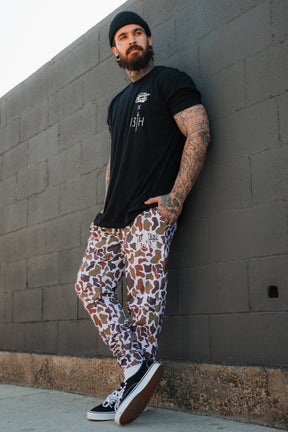 A bearded man in the "Lift Heavy. Pray Hard." black tee by Tango Charlie Apparel, black beanie, camo pants, and black Vans stands against a gray brick wall with one foot crossed, hands in pockets, embodying spiritual resilience.