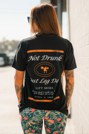 A woman with long hair and tattoos, in sunglasses and colorful leggings, stands outside facing a white wall, wearing the Tango Charlie Apparel "Just Leg Day- Black" workout t-shirt.