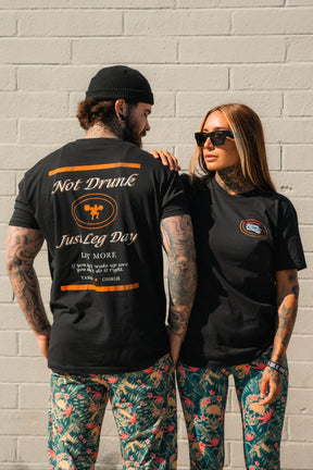 A tattooed man and woman wear matching Just Leg Day- Black shirts by Tango Charlie Apparel. The man shows off the "Not Drunk, Just Leg Day" back text while the woman pairs hers with sunglasses and floral pants for a fun gym look.