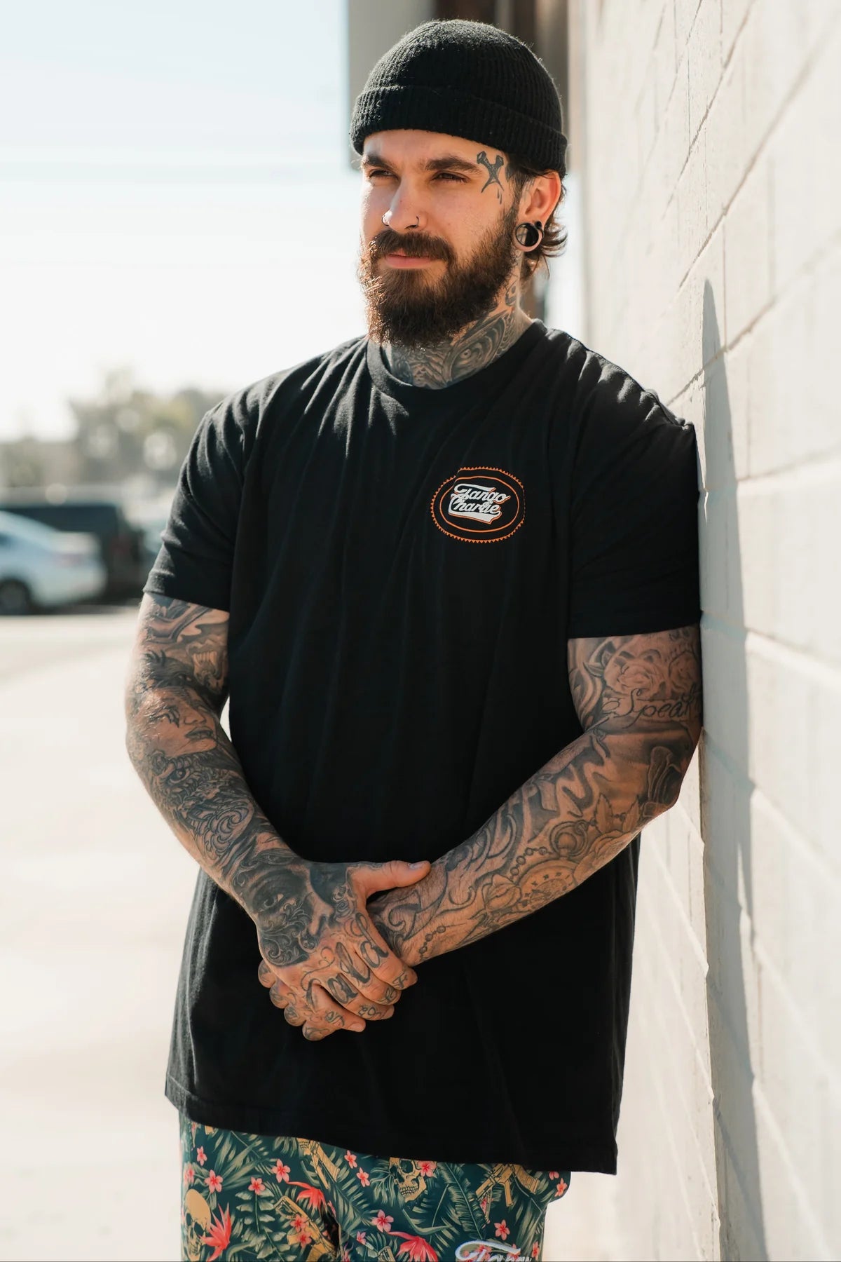 A bearded man with tattoos and a black beanie leans against a white wall, outdoors on a sunny day, wearing the Tango Charlie Apparel Just Leg Day- Black workout t-shirt with a small logo and colorful tropical-patterned shorts.