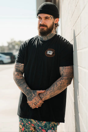 A bearded man with tattoos and a black beanie leans against a white wall, outdoors on a sunny day, wearing the Tango Charlie Apparel Just Leg Day- Black workout t-shirt with a small logo and colorful tropical-patterned shorts.