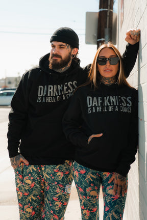 A man and woman stand outdoors in matching Tango Charlie Apparel Darkness Is A Hell Of A Coach hoodies with bold text and vibrant skull-patterned pants; he pairs his with a beanie, she leans on a wall in sunglasses.