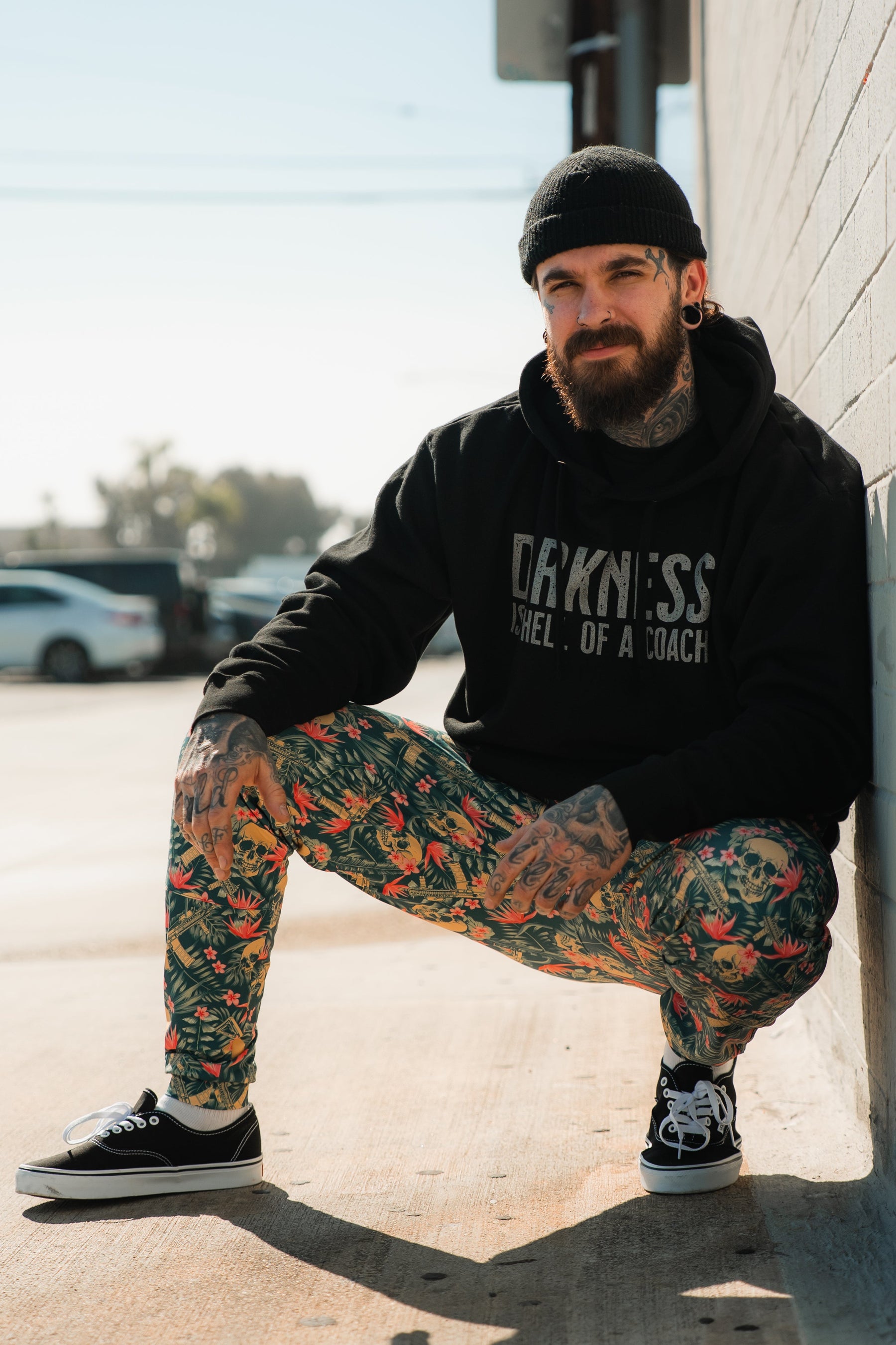 A bearded man with tattoos smiles as he squats outdoors by a wall wearing the Darkness Is A Hell Of A Coach Hoodie from Tango Charlie Apparel, paired with colorful floral pants, a black beanie, and black Vans sneakers. Sunlight casts shadows on the pavement.
