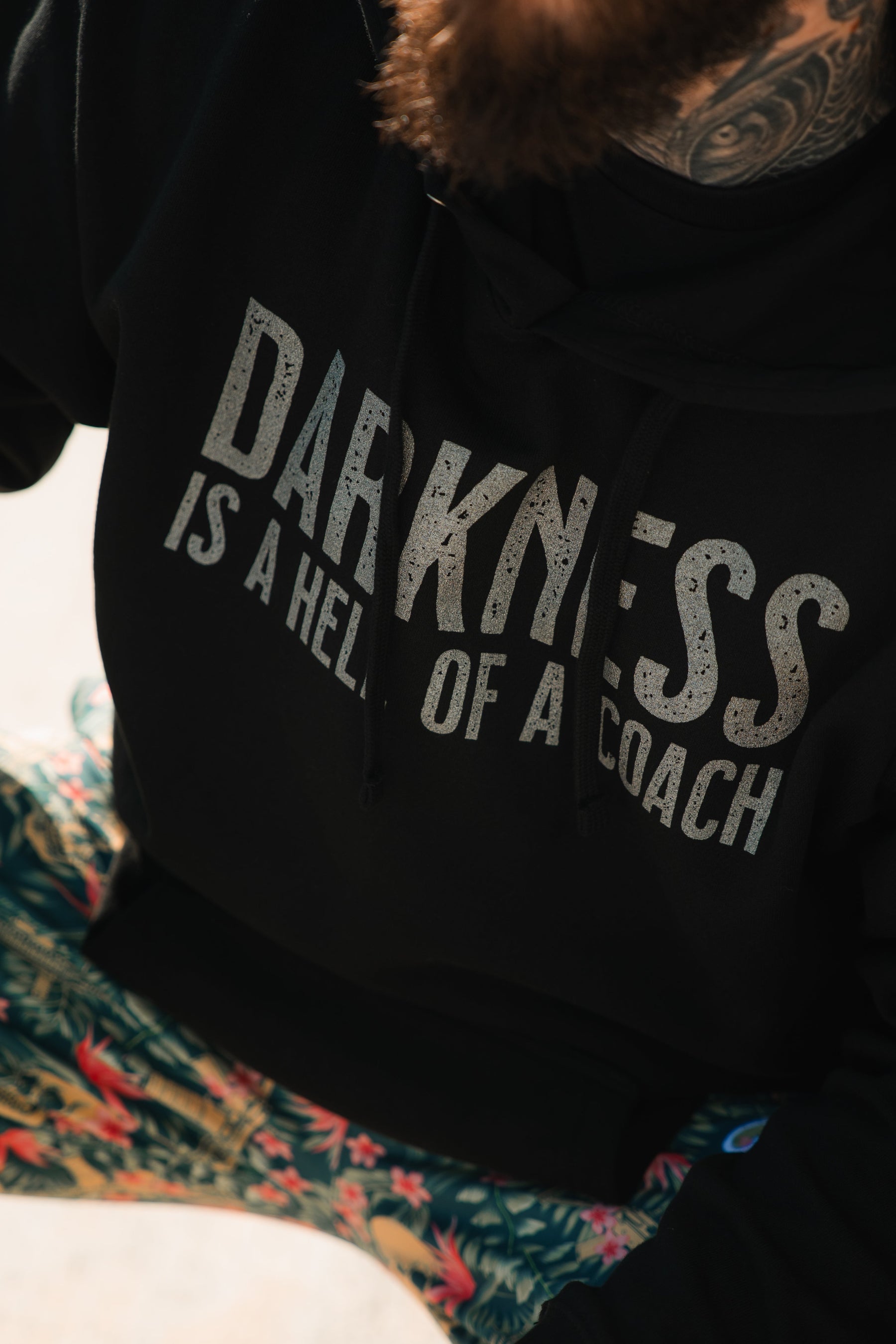 Wearing the Tango Charlie Apparel “Darkness Is A Hell Of A Coach Hoodie,” a person with a beard and neck tattoo sits in colorful gym pants, embodying victory over darkness.
