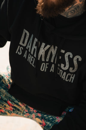 Wearing the Tango Charlie Apparel “Darkness Is A Hell Of A Coach Hoodie,” a person with a beard and neck tattoo sits in colorful gym pants, embodying victory over darkness.