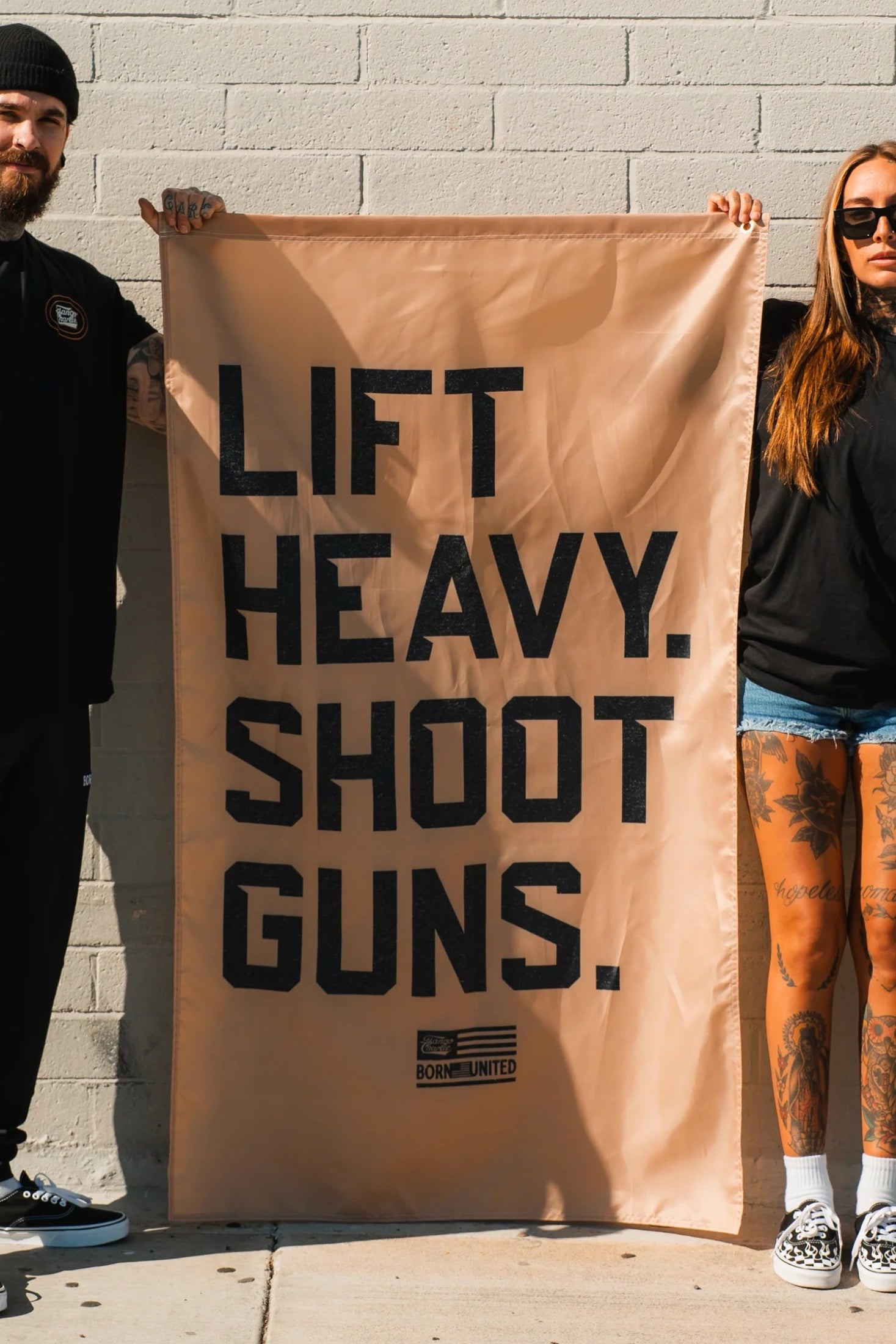 A tattooed man and woman in black shirts and sunglasses stand against a brick wall, holding the Lift Heavy. Shoot Guns. Flag from Tango Charlie Apparel, looking directly at the camera.