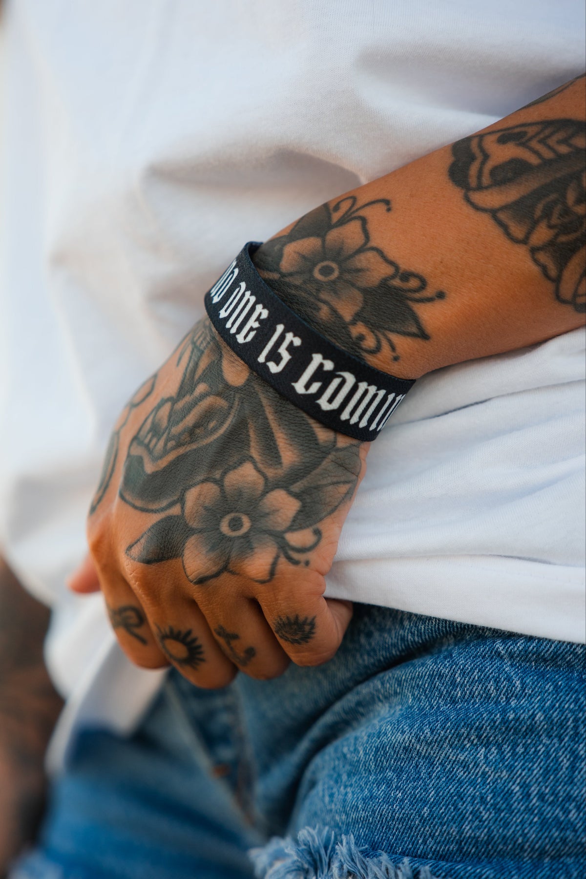 A tattooed hand rests on denim shorts, wearing a white shirt and the "No One Is Coming To Save You" wristband by Tango Charlie Apparel.