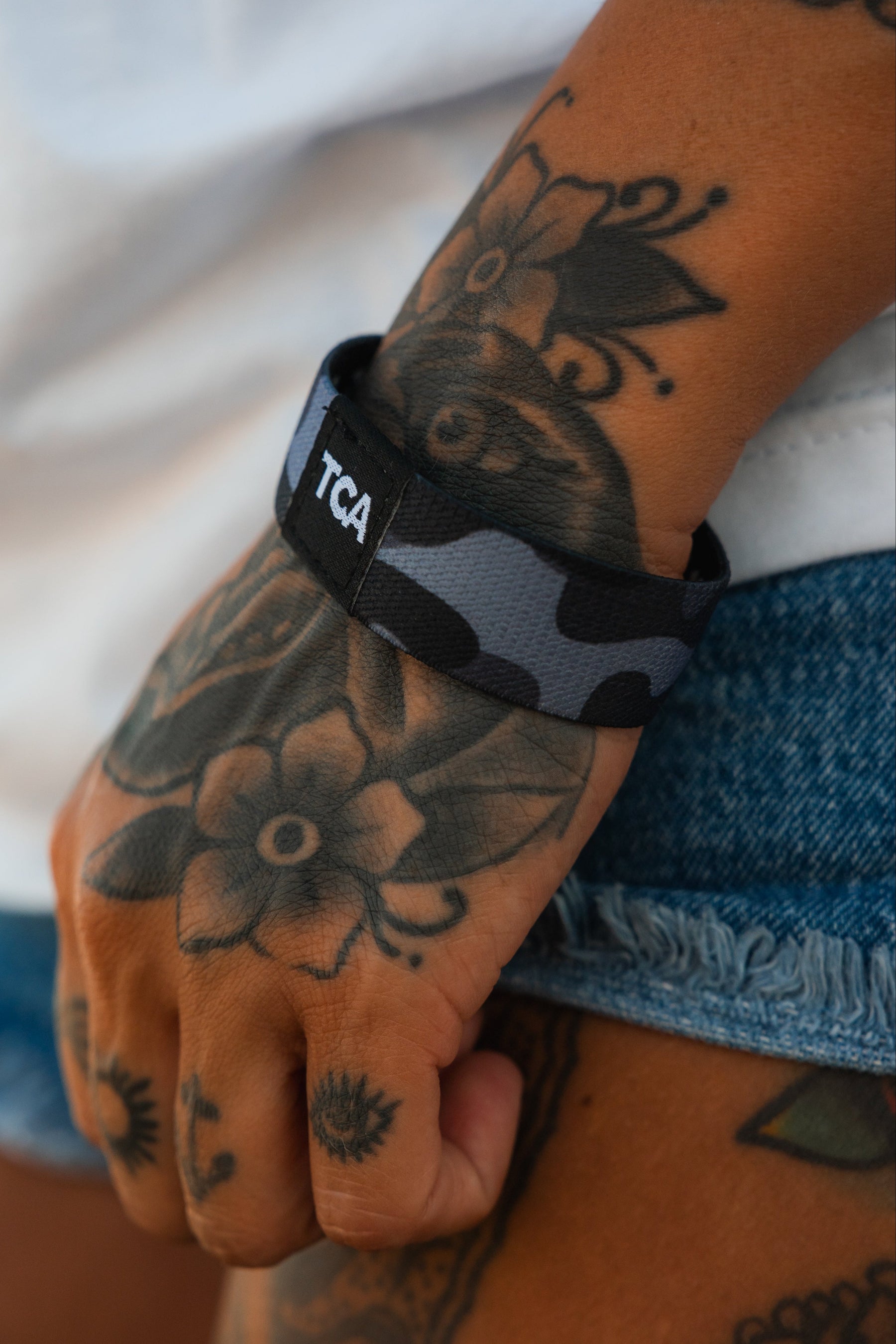 A tattooed hand and wrist wear the Tango Charlie Apparel "No One Is Coming To Save You" wristband, black and gray camouflage-patterned, with denim shorts and a white shirt partially visible.
