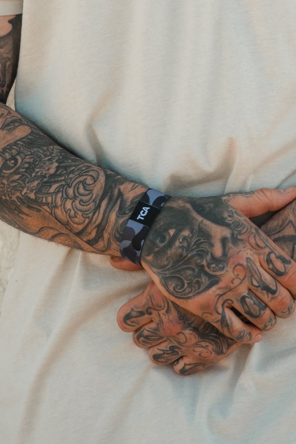 A person with heavily tattooed arms, wearing a short-sleeved white shirt and a black camo Tango Charlie Apparel "No One Is Coming To Save You" wristband marked with TCA.