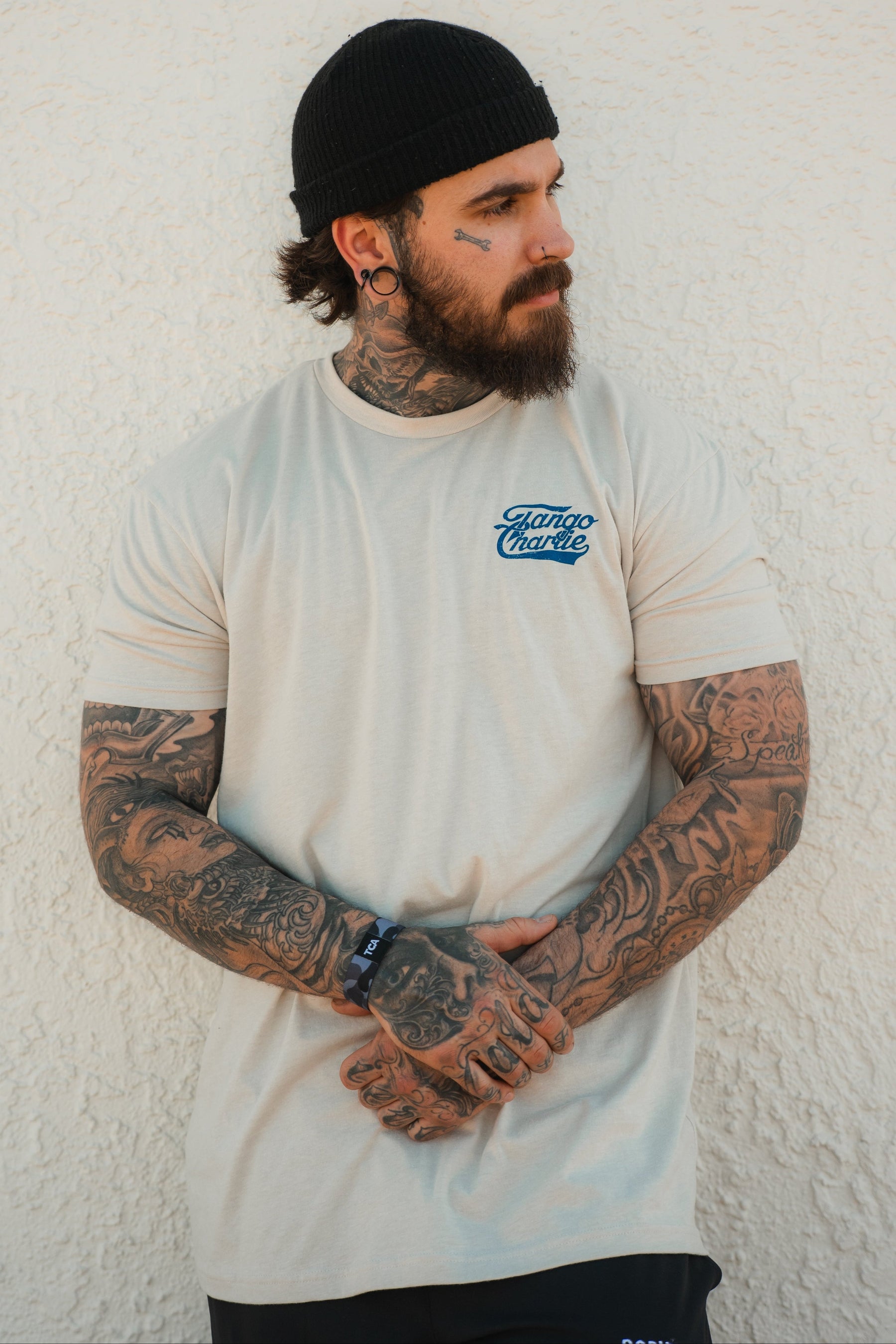 A bearded man with arm and face tattoos stands against a textured white wall, wearing the Tango Charlie Apparel "Foundational Five - Tee," a beige shirt reading "Faith Family Freedom," along with a black beanie and shorts, hands clasped in front.