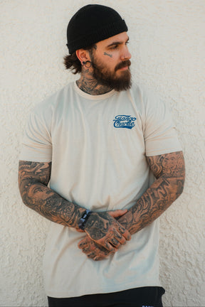 A bearded man with arm and face tattoos stands against a textured white wall, wearing the Tango Charlie Apparel "Foundational Five - Tee," a beige shirt reading "Faith Family Freedom," along with a black beanie and shorts, hands clasped in front.
