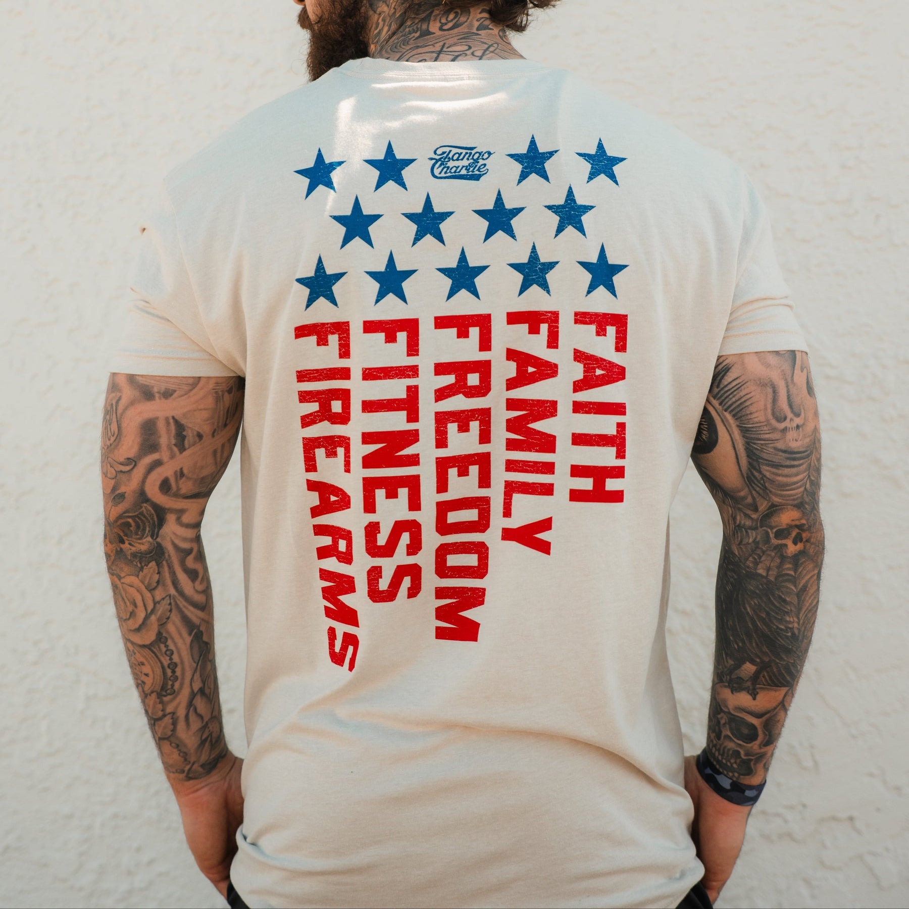 A tattooed person wears the Tango Charlie Apparel Foundational Five - Tee, a beige shirt with blue stars and bold red "Faith Family Freedom, Fitness, Firearms" text on the back.