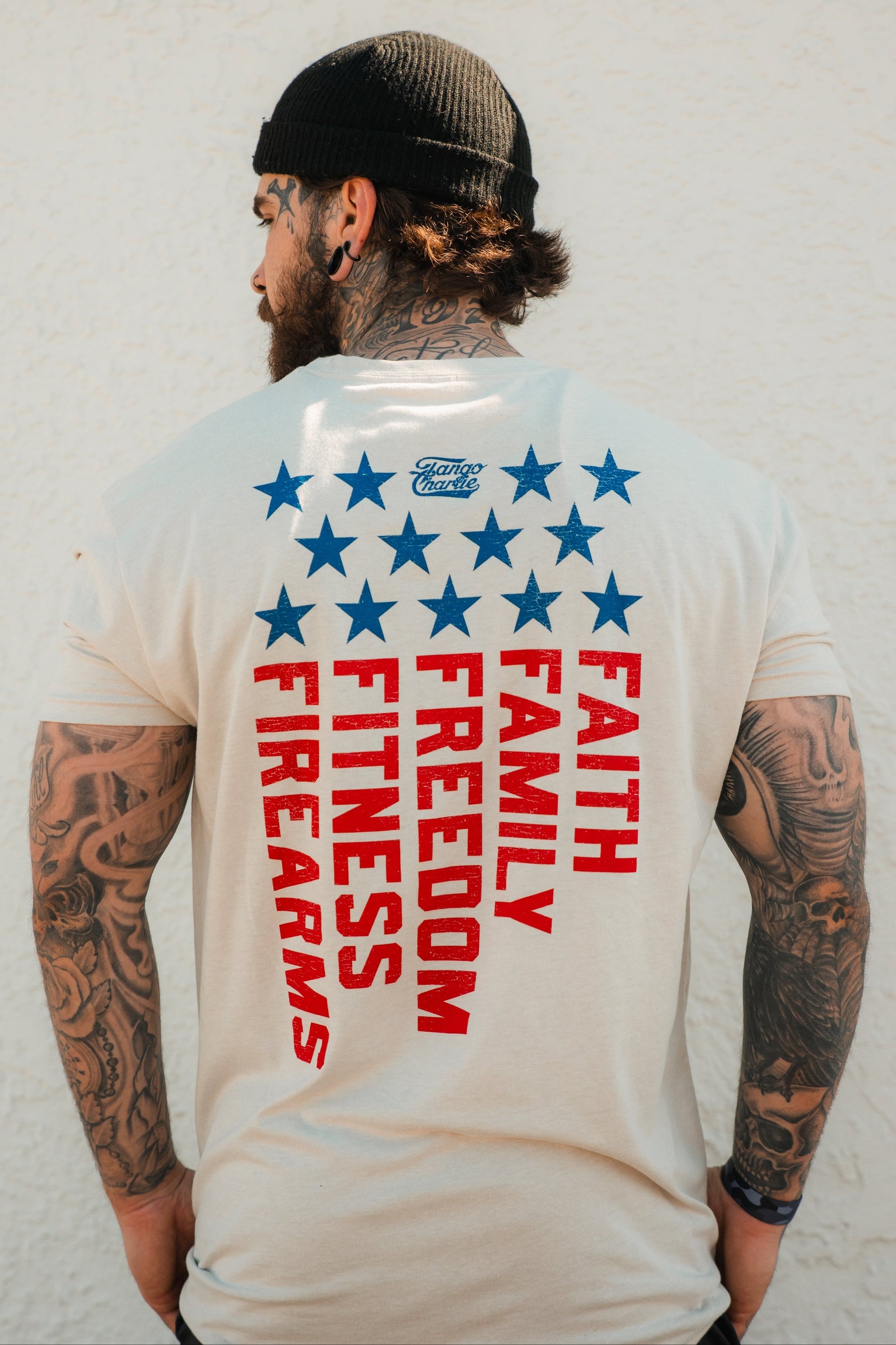A tattooed man in a black beanie and the Tango Charlie Apparel Foundational Five - Tee, featuring “Faith Family Freedom” in bold red, stands before a textured white wall, showcasing pride in his values.