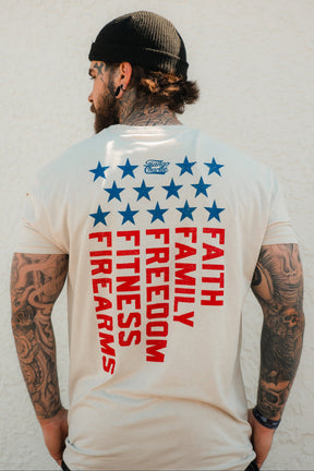 A tattooed man in a black beanie and the Tango Charlie Apparel Foundational Five - Tee, featuring “Faith Family Freedom” in bold red, stands before a textured white wall, showcasing pride in his values.