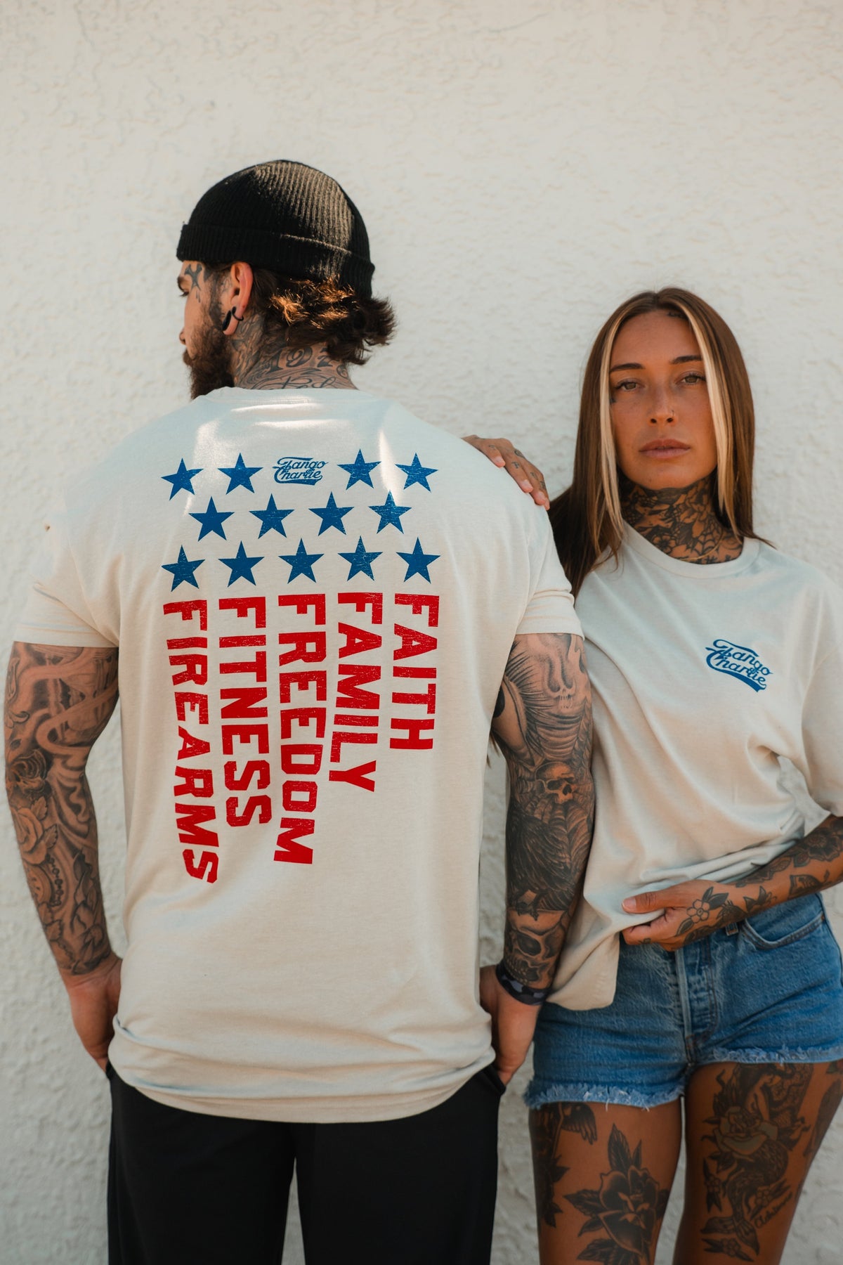 Two tattooed people pose against a white wall. The man faces away, showing off a Tango Charlie Apparel Foundational Five - Tee with blue stars and bold red words: Faith Family Freedom. The woman stands in front, wearing the matching tee and denim shorts.