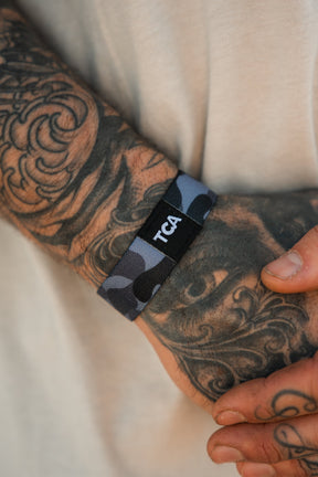 A close-up of a tattooed hand wearing the Tango Charlie Apparel "No One Is Coming To Save You" wristband, featuring a black and grey pattern with TCA lettering, set against a blurred light background.