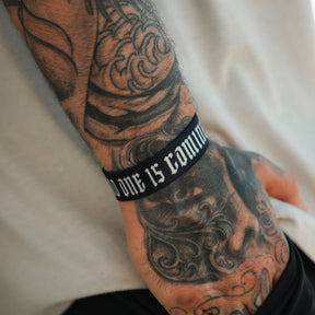 A tattooed hand and wrist wears a Tango Charlie Apparel "No One Is Coming To Save You" black motivational wristband with white text, paired with a light-colored shirt.