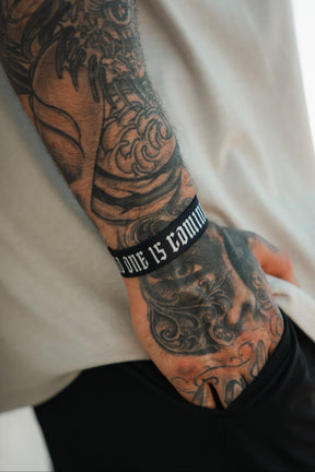 A tattooed arm with bold designs rests in a pocket, wearing Tango Charlie Apparel’s "No One Is Coming To Save You" black wristband featuring white Gothic-style text.