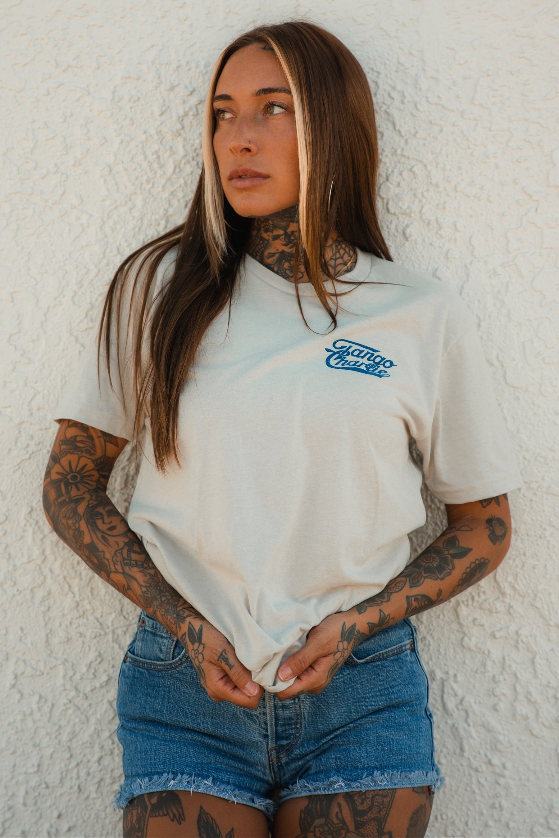 A woman with long brown hair and tattoos stands against a textured white wall, wearing the "Foundational Five - Tee" by Tango Charlie Apparel paired with denim shorts, looking to the side while holding her shirt.