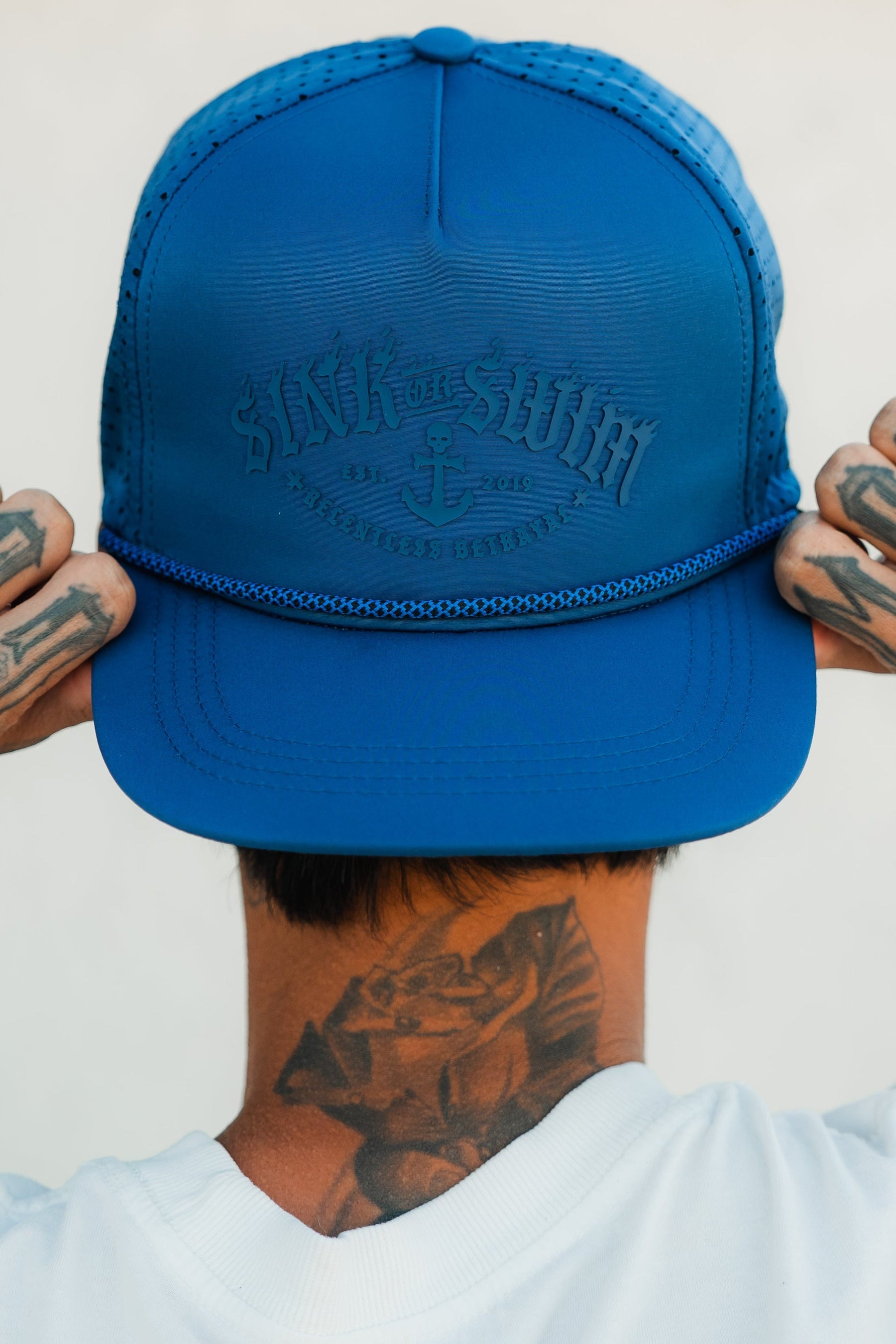 A person wearing a Dark Waters SOS Mute PVC Snapback by Relentless Betrayal is seen from behind, holding the hat with both hands. With tattoos on their hands and neck, they embody a pirate lifestyle while wearing a white shirt.