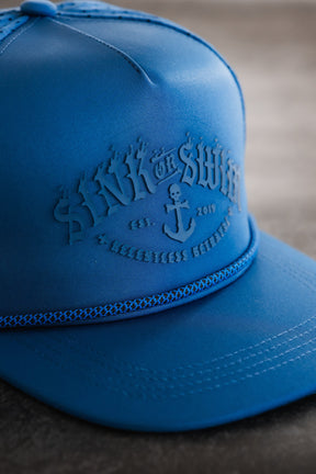 A close-up of the Relentless Betrayal Dark Waters SOS Mute PVC Snapback shows a blue patch embossed with SINK OR SWIM, an anchor, EST. 2019, and RELENTLESS PURSUIT—pirate-inspired style with a braided band above the brim.