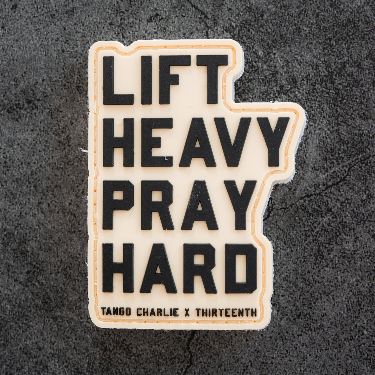 The Lift Heavy. Pray Hard. - Patch by Tango Charlie Apparel features bold black 3D text on a textured dark gray 3-inch PVC patch with Velcro backing—perfect for your gear.