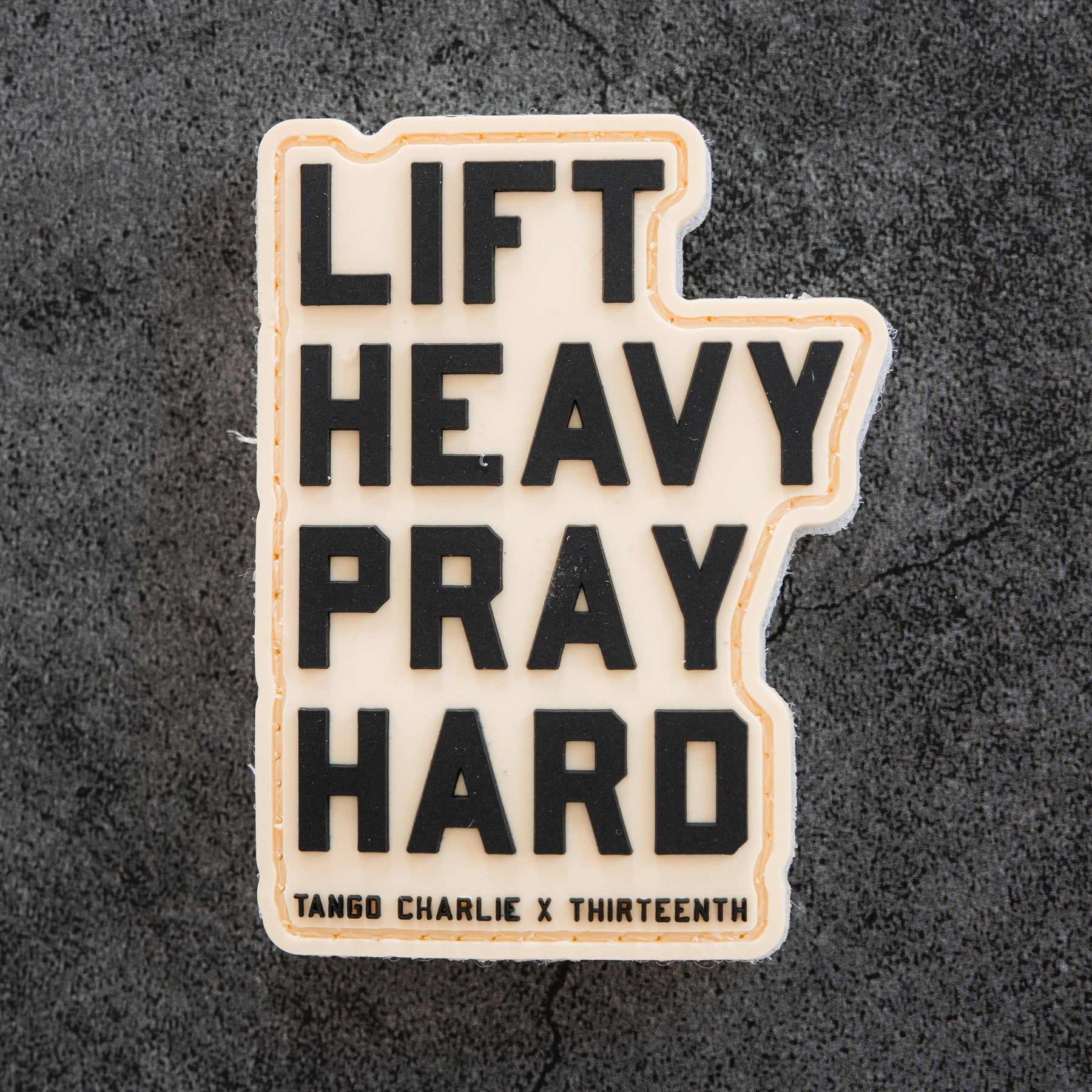 The Lift Heavy. Pray Hard. - Patch by Tango Charlie Apparel features bold black 3D text on a textured dark gray 3-inch PVC patch with Velcro backing—perfect for your gear.