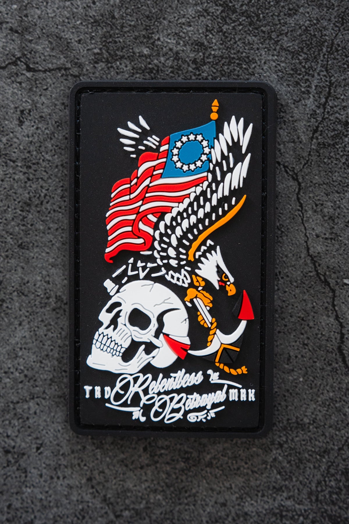 The Revolution Patch by Relentless Betrayal is a 3x3" 3D PVC patch featuring a skull, outstretched eagle, and stylized American flag, with "The Relentless Betrayal" below. It’s shown on a rough, dark surface.