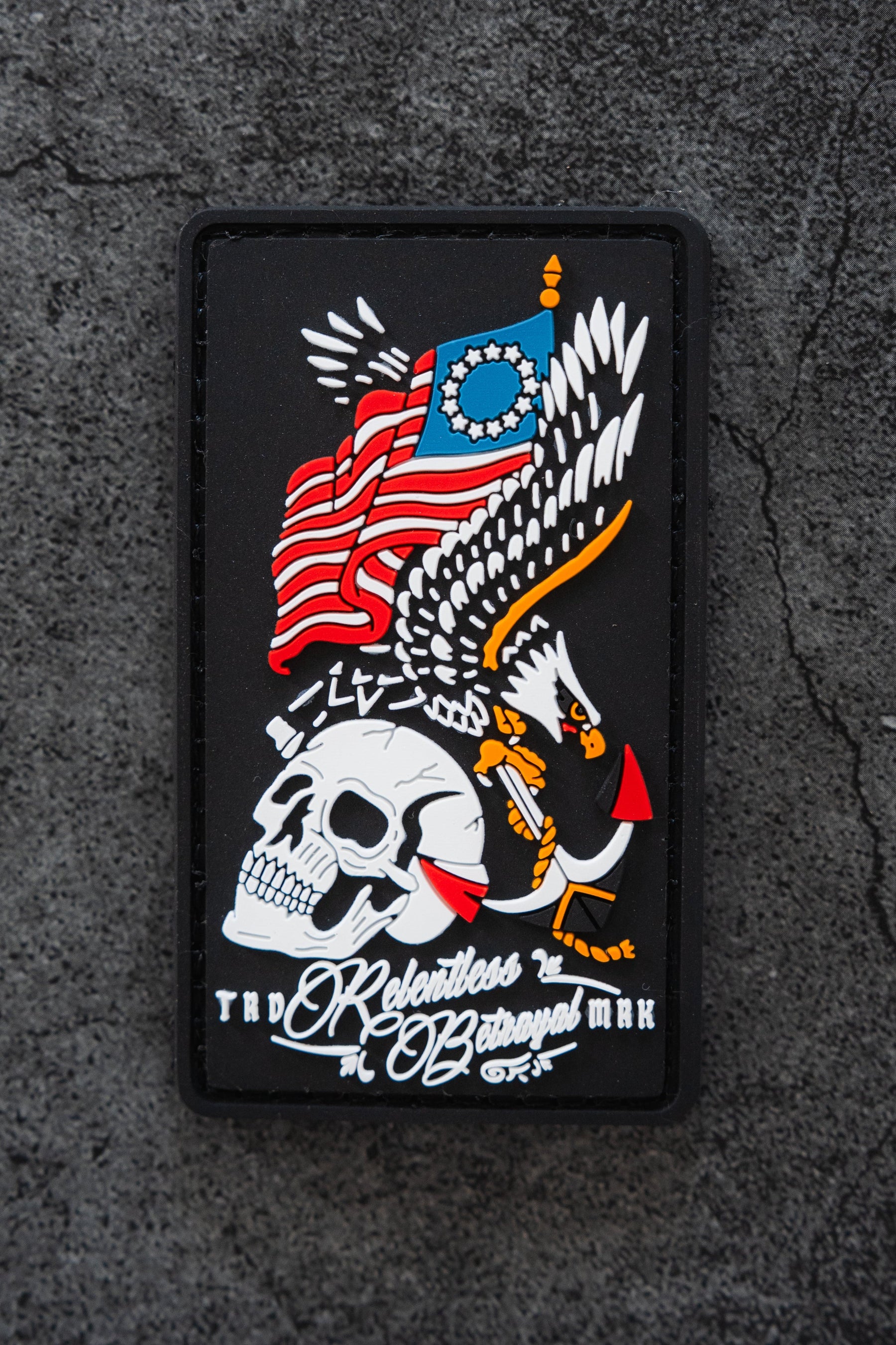 The Revolution Patch by Relentless Betrayal is a 3x3" 3D PVC patch featuring a skull, outstretched eagle, and stylized American flag, with "The Relentless Betrayal" below. It’s shown on a rough, dark surface.