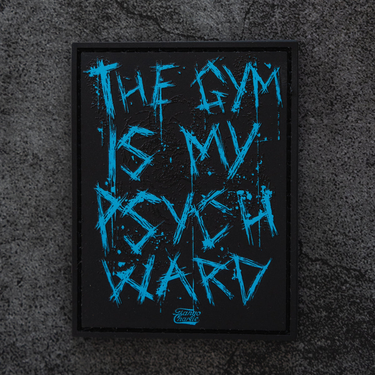 The Psych Ward Patch by Tango Charlie Apparel is a black rectangular Velcro patch with bold blue jagged text reading “THE GYM IS MY PSYCH WARD” on a rugged dark textured background.