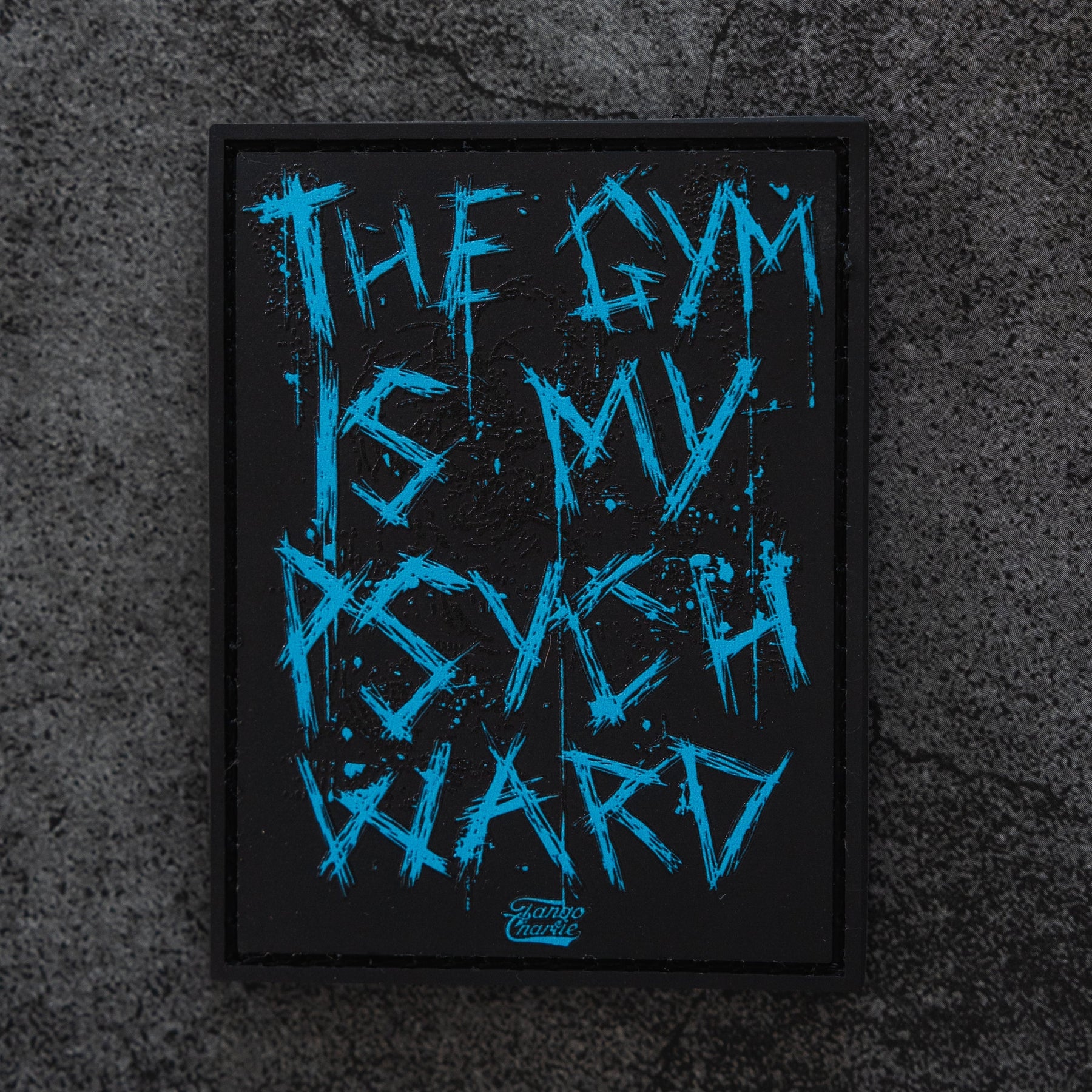 The Psych Ward Patch by Tango Charlie Apparel is a black rectangular Velcro patch with bold blue jagged text reading “THE GYM IS MY PSYCH WARD” on a rugged dark textured background.