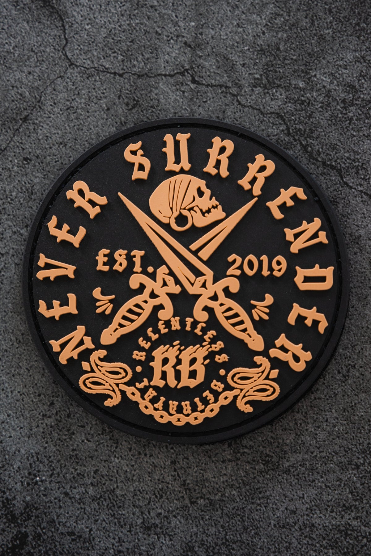 The Never Surrender Patch by Relentless Betrayal is a black 3x3" round PVC Velcro patch with orange text, skull above scissors, EST. 2019, and KB SELECTED QUALITY at the bottom, set on a rough stone background.