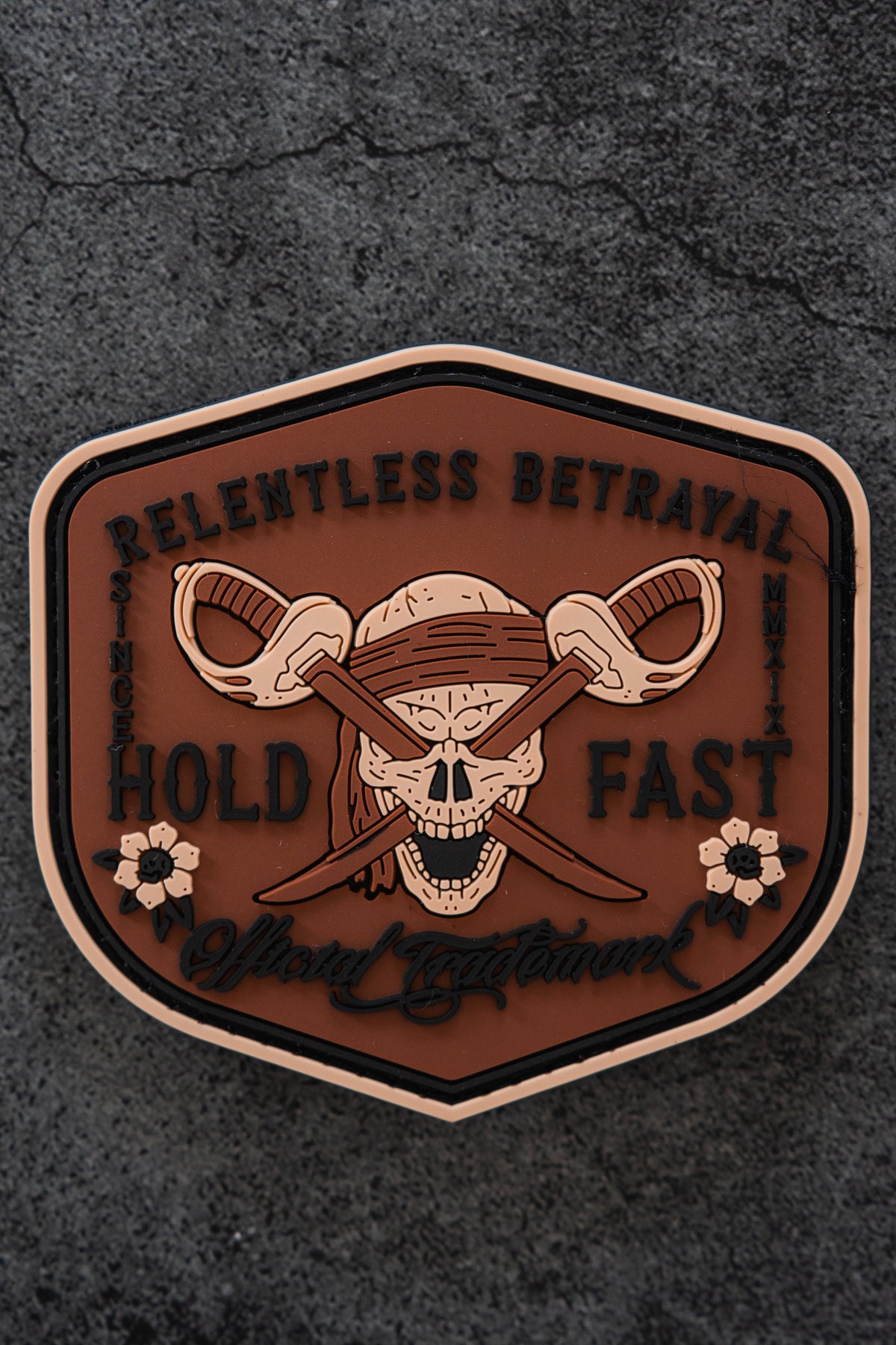 The Hold Fast Patch by Relentless Betrayal is a 3-inch Velcro patch featuring a skull with a bandana and crossed swords, surrounded by "Relentless Betrayal, Hold Fast," floral accents, and set on a dark textured background.