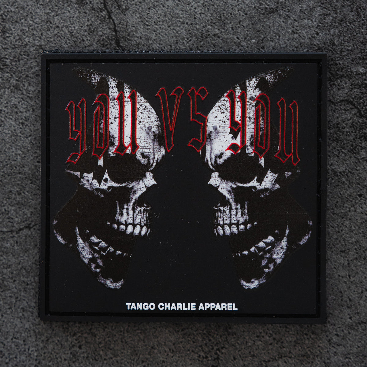 The You vs You - Patch by Tango Charlie Apparel is a black 3D PVC patch with two mirrored skulls, red "you vs you" text above, and Tango Charlie Apparel in white at the bottom, displayed on a concrete background.