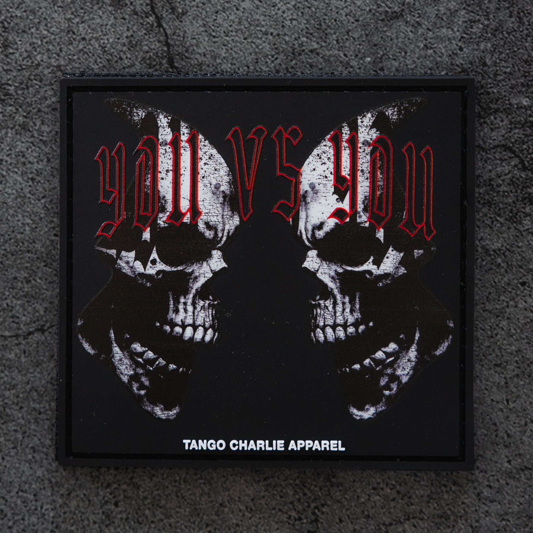 The You vs You - Patch by Tango Charlie Apparel is a black 3D PVC patch with two mirrored skulls, red "you vs you" text above, and Tango Charlie Apparel in white at the bottom, displayed on a concrete background.