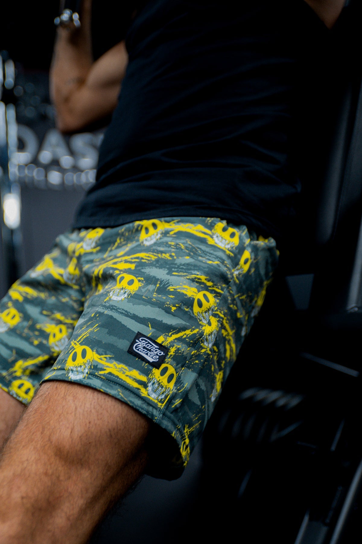 Someone wearing Tango Charlie Apparel’s Men's Performance Training Shorts | 5.5"- Savage in green camo with yellow skulls is working out on gym equipment; only their lower body and a bit of a black shirt are visible.