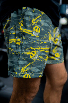 Close-up of a person wearing Tango Charlie Apparel Men's Performance Training Shorts | 5.5"- Savage in green, featuring yellow smiley face graphics and splatter patterns. The person’s legs and part of a black shirt are visible.