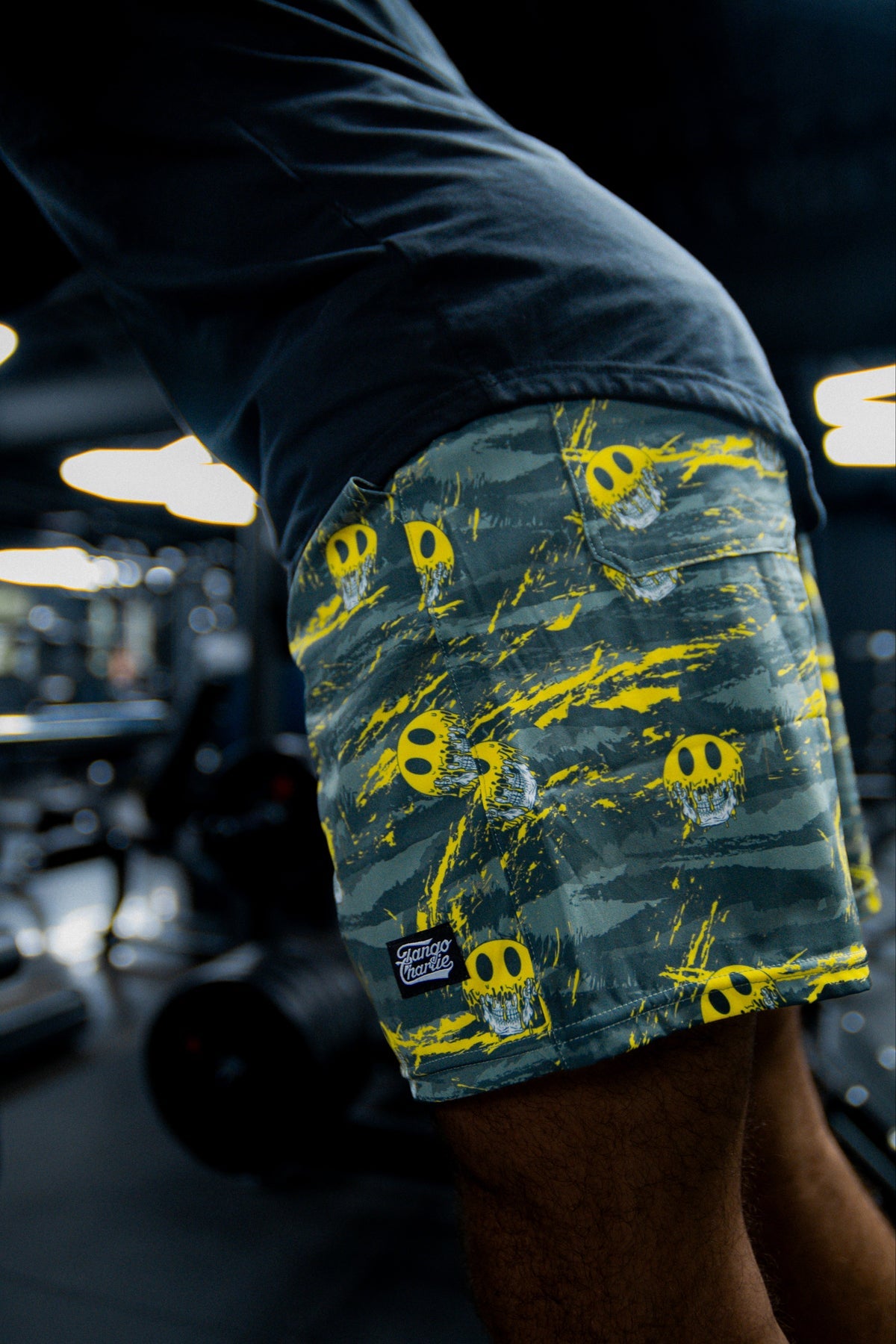 A person wearing Tango Charlie Apparel Men's Performance Training Shorts | 5.5"- Savage, featuring yellow skull patterns, is working out in a gym with gym equipment visible in the blurred background.