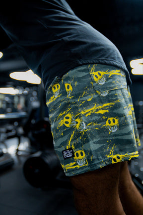 A person wearing Tango Charlie Apparel Men's Performance Training Shorts | 5.5"- Savage, featuring yellow skull patterns, is working out in a gym with gym equipment visible in the blurred background.