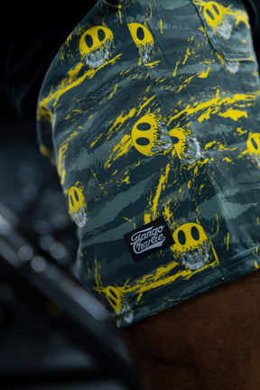 Close-up of a person wearing Men's Performance Training Shorts | 5.5"- Savage by Tango Charlie Apparel, with a green and black pattern, yellow smiley face skulls, and a Tango Charlie brand patch on the pocket.