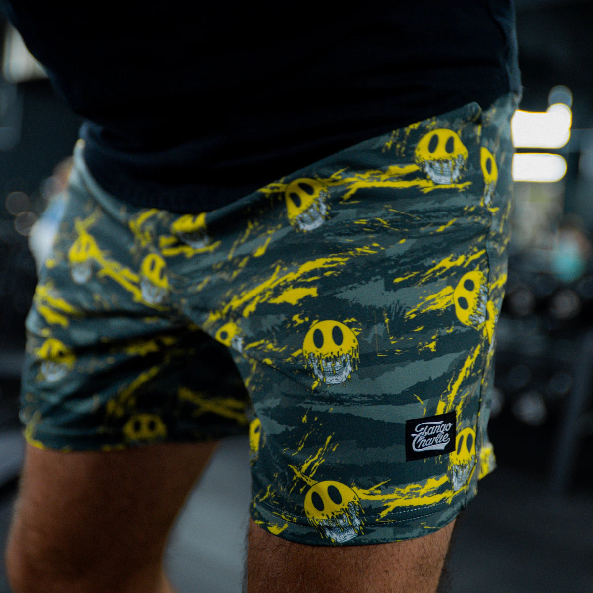 A person wears Tango Charlie Apparel Men's Performance Training Shorts | 5.5"- Savage, featuring green camo with bright yellow skulls, indoors with blurred gym equipment in the background.