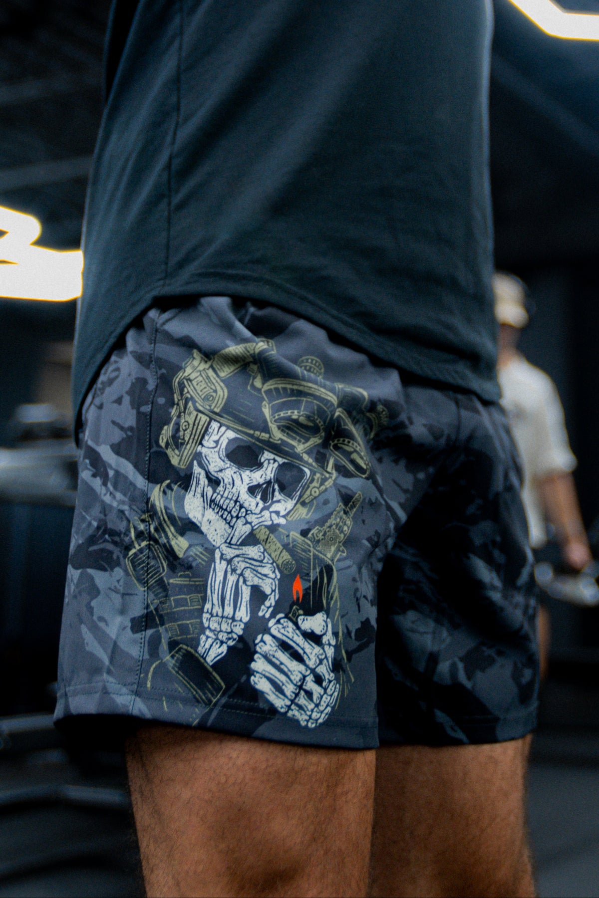 A person wears Born United Private Labels Mens Performance Hybrid Shorts 5.5 | Savage Ops, featuring a skeleton in tactical gear holding a lighter with a red flame; the blurred background highlights the unique design.