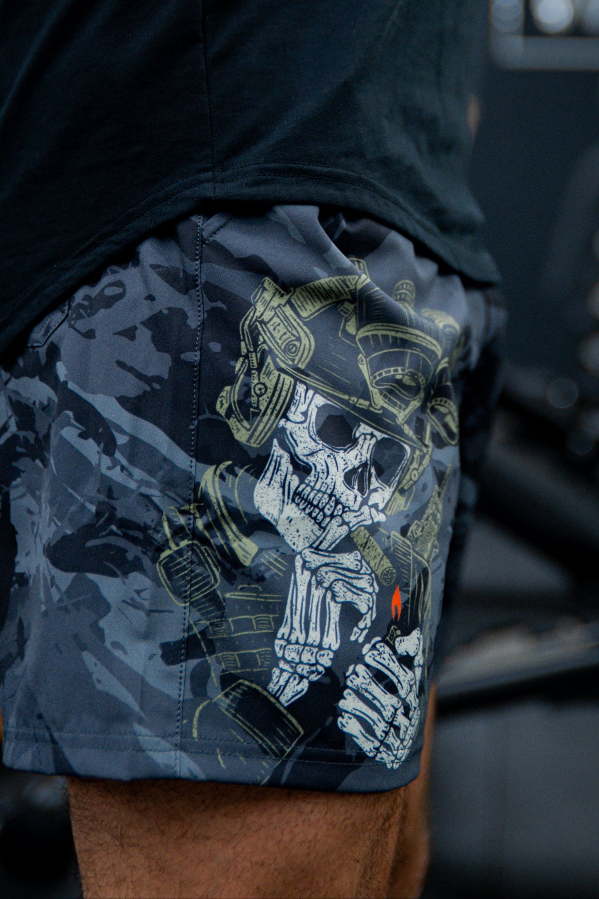 A close-up of a person wearing Born United Private Label Mens Performance Hybrid Shorts 5.5 | Savage Ops, which feature a detailed graphic of a skeleton soldier in a helmet lighting a cigarette with a visible red flame.