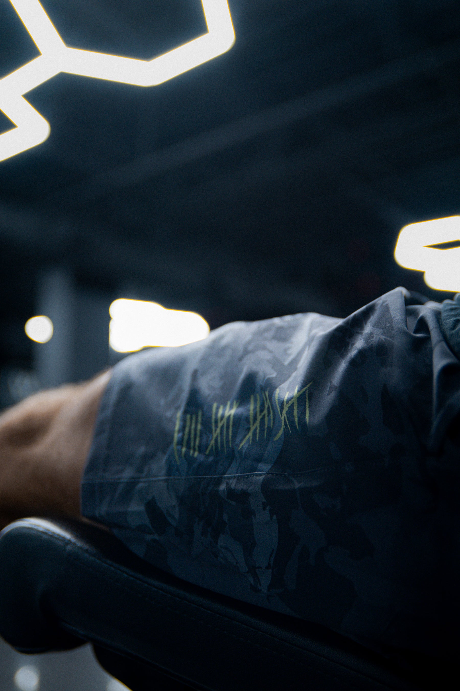 A close-up of a leg wearing Born United Private Label Mens Performance Hybrid Shorts 5.5 | Savage Ops in dark camo with green text, made from hybrid materials, set in a gym with geometric ceiling lights behind.