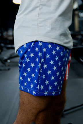 A person wears Born United Private Label Mens Performance Hybrid Shorts 5.5 | Stars & Stripes, featuring a blue fabric with white stars. Only their lower torso and upper legs are visible, with blurred gym equipment in the background.