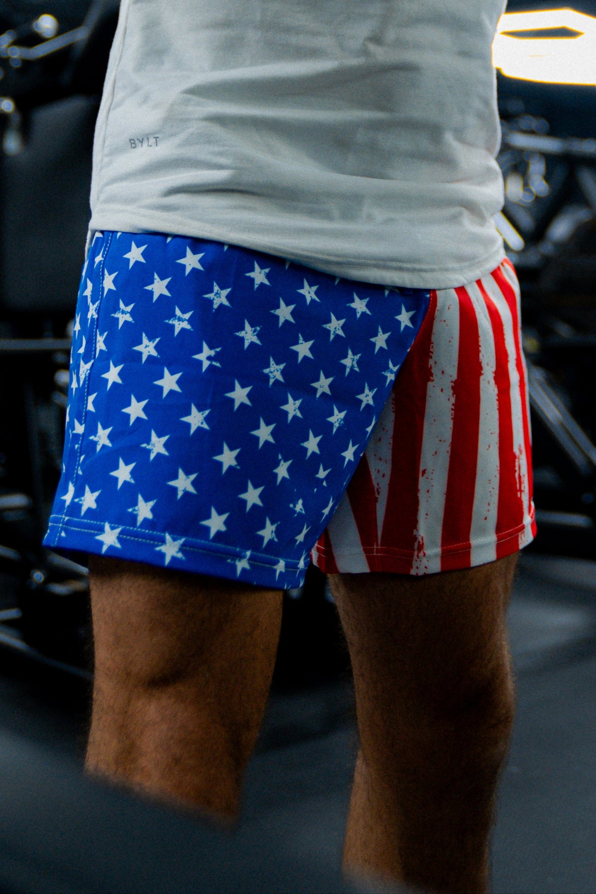 A person stands indoors, partially visible from the waist down, wearing Born United Private Label Mens Performance Hybrid Shorts 5.5 | Stars & Stripes—blue with white stars on one side, red and white stripes on the other.