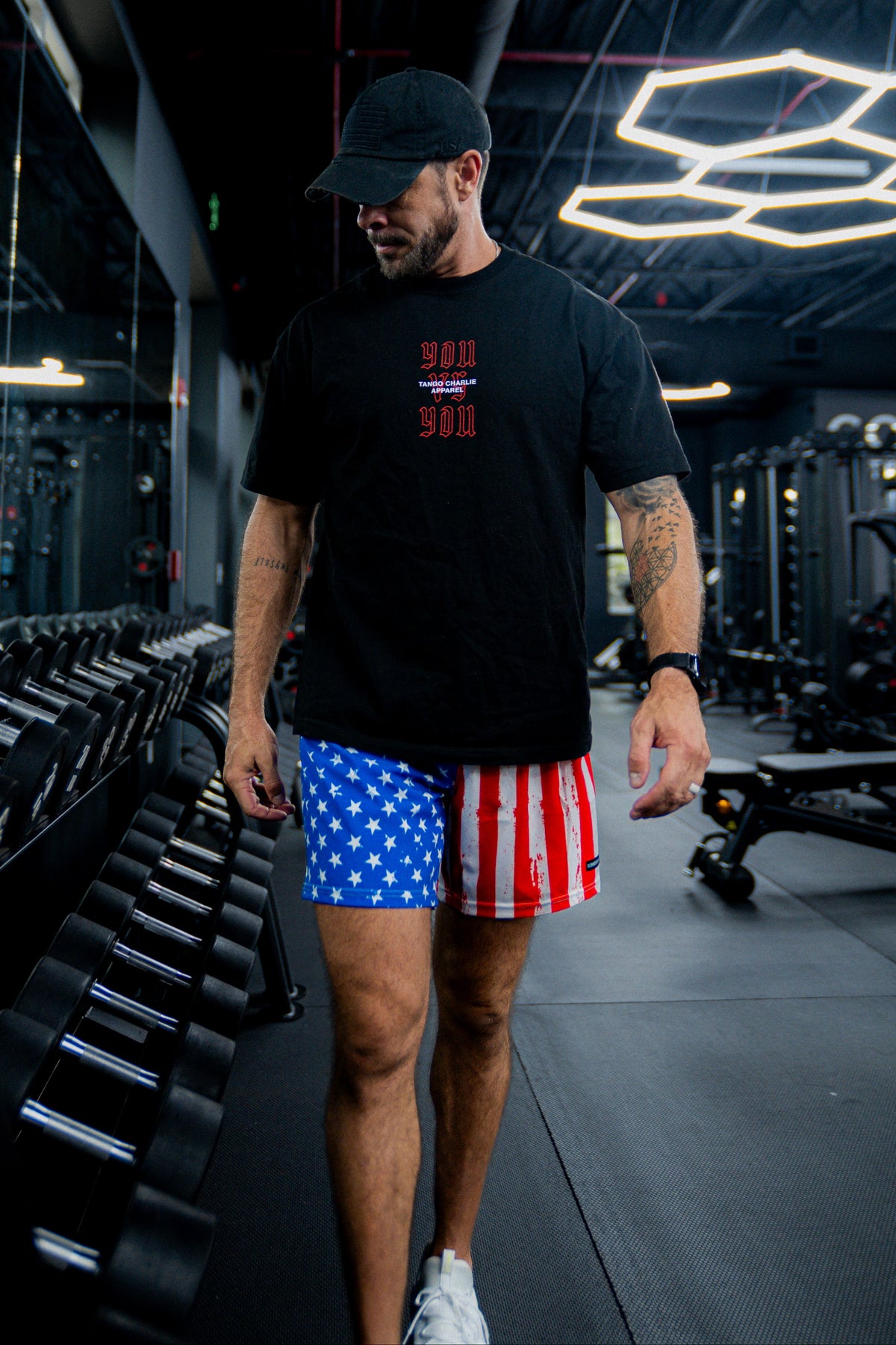 A man in a black cap, black t-shirt, and Born United Private Label Mens Performance Hybrid Shorts 5.5 | Stars & Stripes walks through a sleek, modern gym filled with dumbbells and workout equipment under stylish lighting.