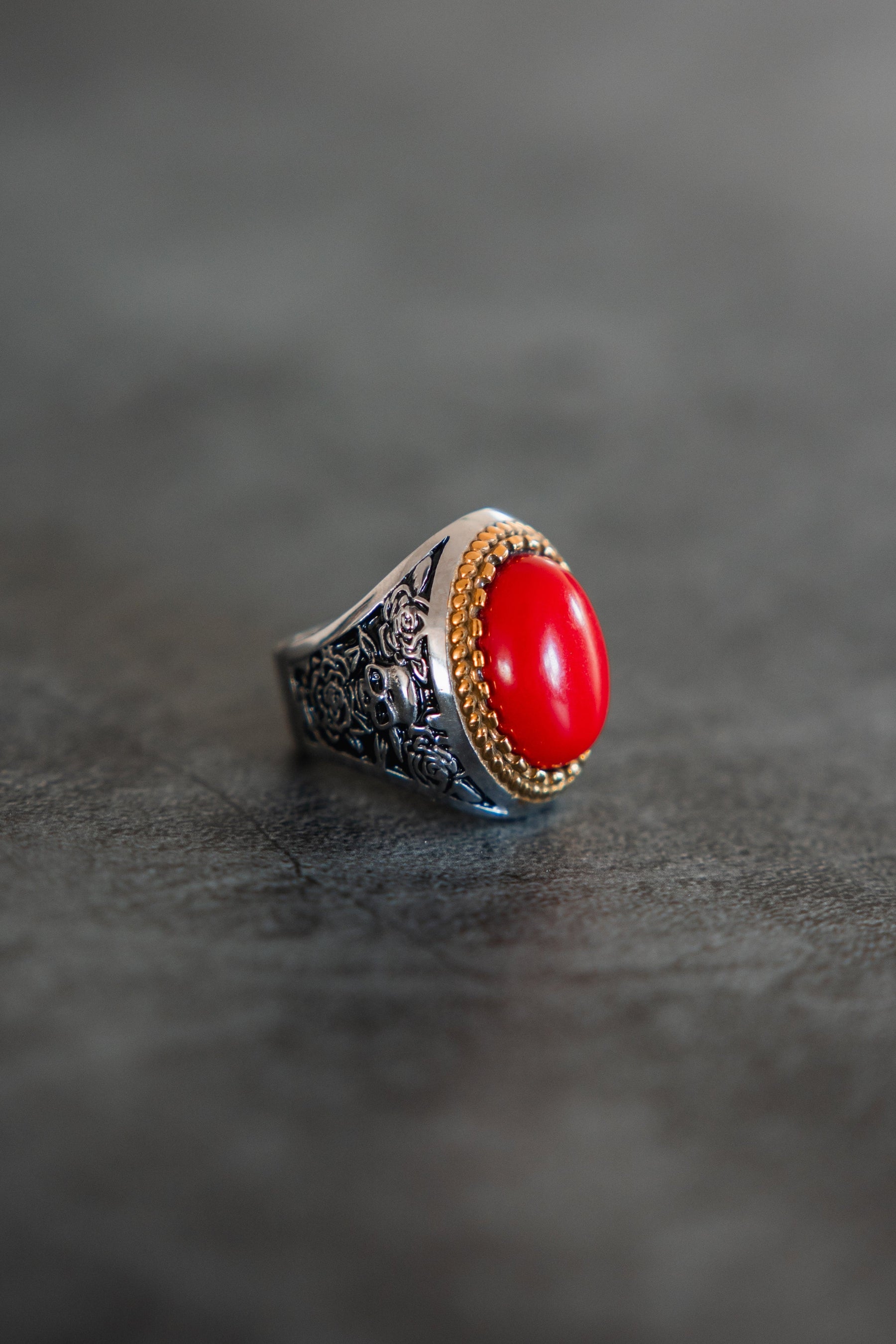 The Burning Rose Ring by Relentless Betrayal features ornate floral engravings and a large oval red stone in a gold bezel, showcased on a gray surface.